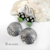 These handcrafted silver pewter earrings have a hand stamped texture and are accented with artisan handmade lampwork beads, green serpentine gemstones and off-white faceted crystal beads. The lampwork beads have a black base with off-white dots that have subtle pale yellow undertones, along with funky black bumps sticking out