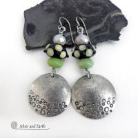 These handcrafted silver pewter earrings have a hand stamped texture and are accented with artisan handmade lampwork beads, green serpentine gemstones and off-white faceted crystal beads. The lampwork beads have a black base with off-white dots that have subtle pale yellow undertones, along with funky black bumps sticking out. Handmade in the USA by Silver and Earth. 
