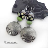 These handcrafted silver pewter earrings have a hand stamped texture and are accented with artisan handmade lampwork beads, green serpentine gemstones and off-white faceted crystal beads. The lampwork beads have a black base with off-white dots that have subtle pale yellow undertones, along with funky black bumps sticking out. Handmade in the USA by Silver and Earth. 