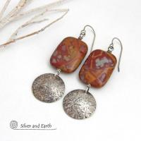 These one-of-a-kind handcrafted earrings feature natural Noreena jasper gemstones accented with textured sterling silver dangles. This stone is known for its dramatic abstract and geometric patterns and is found only in the Pilbara region of Western Australia. The stones in this pair have a great pattern consisting of hues of deep burgundy-red with fiery orange and gray streaks. 