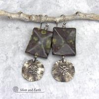 Large chunky spotted jasper stones are accented with handcrafted rustic sterling silver dangles in these one-of-a-kind earrings. The silver has an earthy hammered organic texture with a few holes for a primal effect. The stones have a dark brownish-black base with a natural matrix of green spots and a faceted finish.