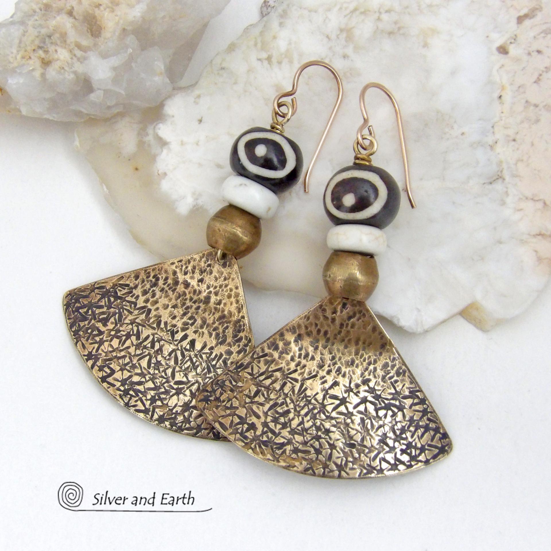 These handcrafted earrings feature gold brass fan-shaped dangles with a rustic hammered texture accented with African batik bone beads, off-white magnesite stones and African brass beads. Unique and striking, they have a bold ethnic vibe with African tribal flair that makes a striking statement. Handmade in the USA by Silver and Earth Jewelry. 