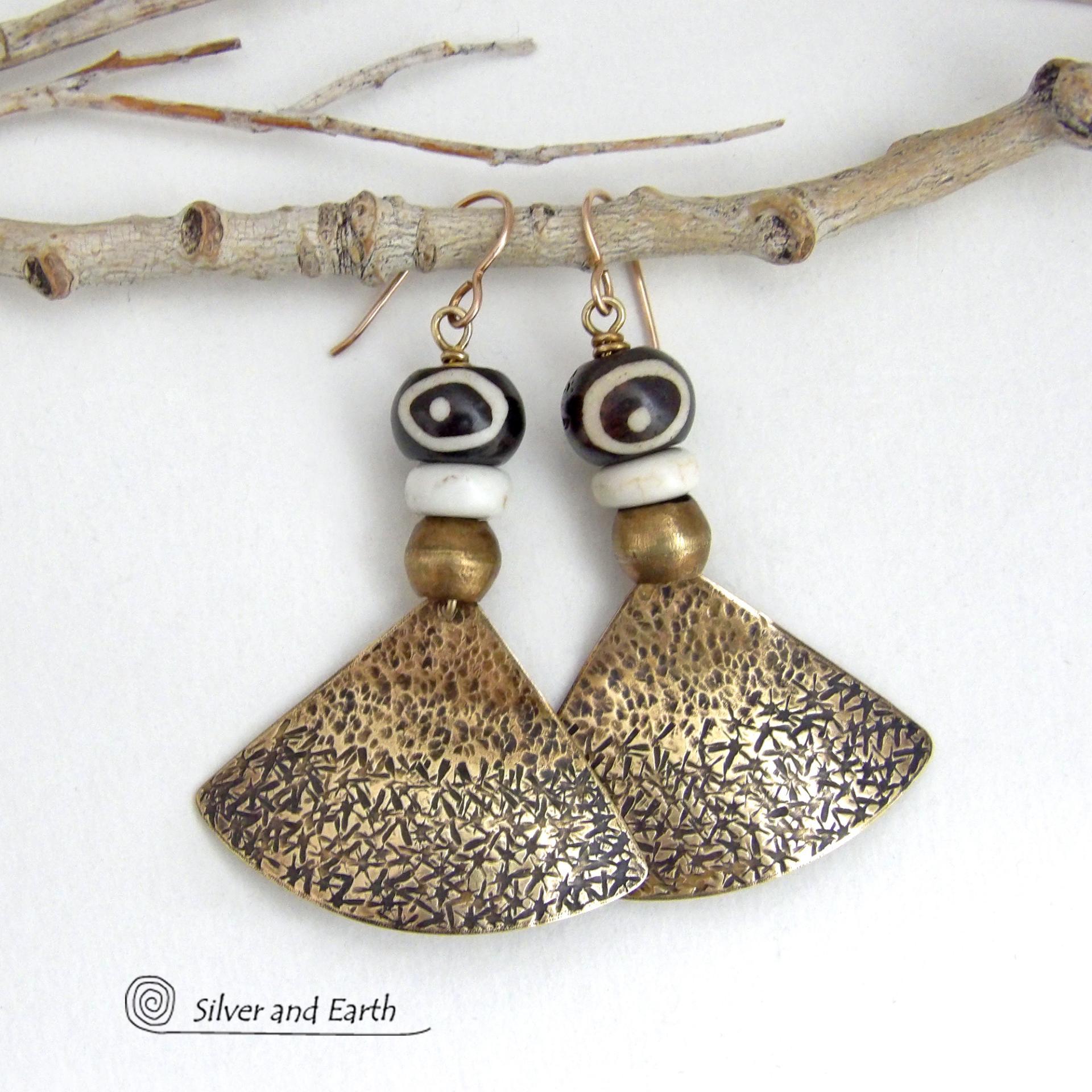 These handcrafted earrings feature gold brass fan-shaped dangles with a rustic hammered texture accented with African batik bone beads, off-white magnesite stones and African brass beads. Unique and striking, they have a bold ethnic vibe with African tribal flair that makes a striking statement. Handmade in the USA by Silver and Earth Jewelry. 