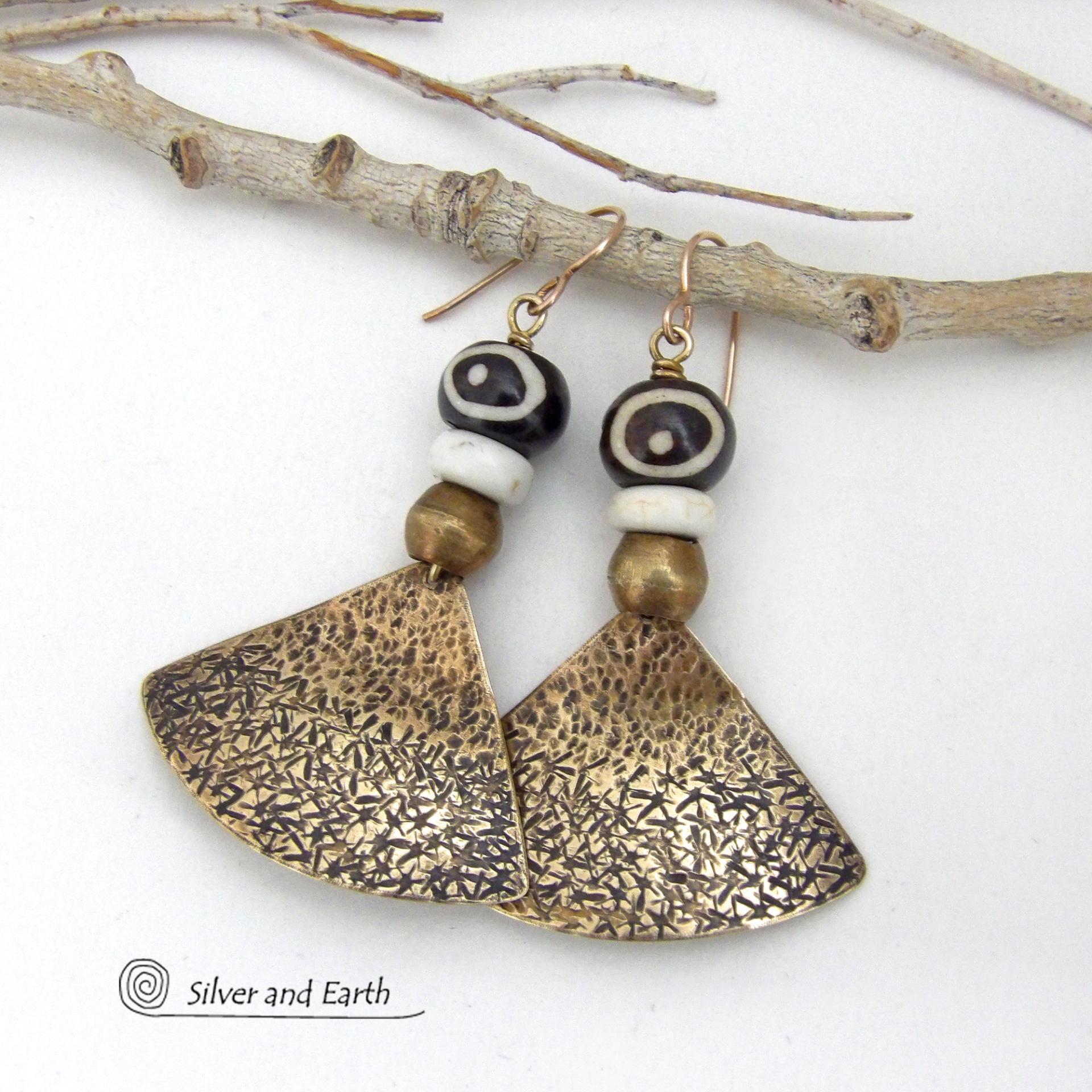 These handcrafted earrings feature gold brass fan-shaped dangles with a rustic hammered texture accented with African batik bone beads, off-white magnesite stones and African brass beads. Unique and striking, they have a bold ethnic vibe with African tribal flair that makes a striking statement. Handmade in the USA by Silver and Earth Jewelry. 