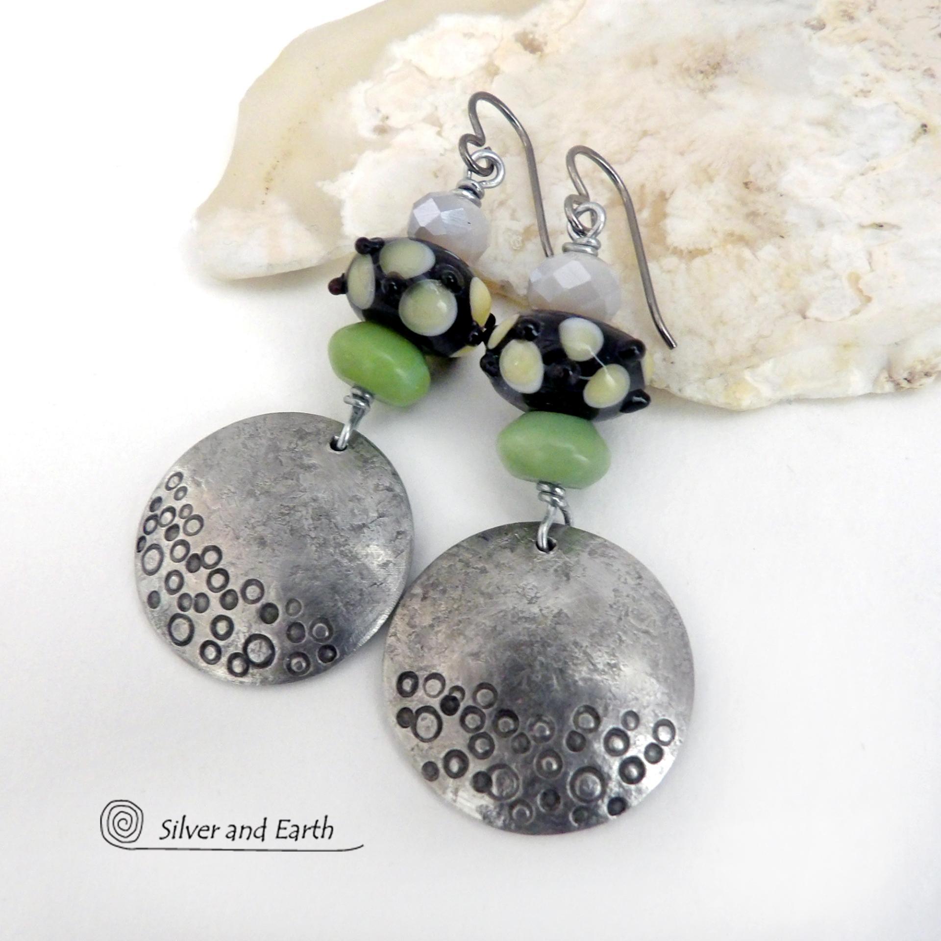 These handcrafted silver pewter earrings have a hand stamped texture and are accented with artisan handmade lampwork beads, green serpentine gemstones and off-white faceted crystal beads. The lampwork beads have a black base with off-white dots that have subtle pale yellow undertones, along with funky black bumps sticking out
