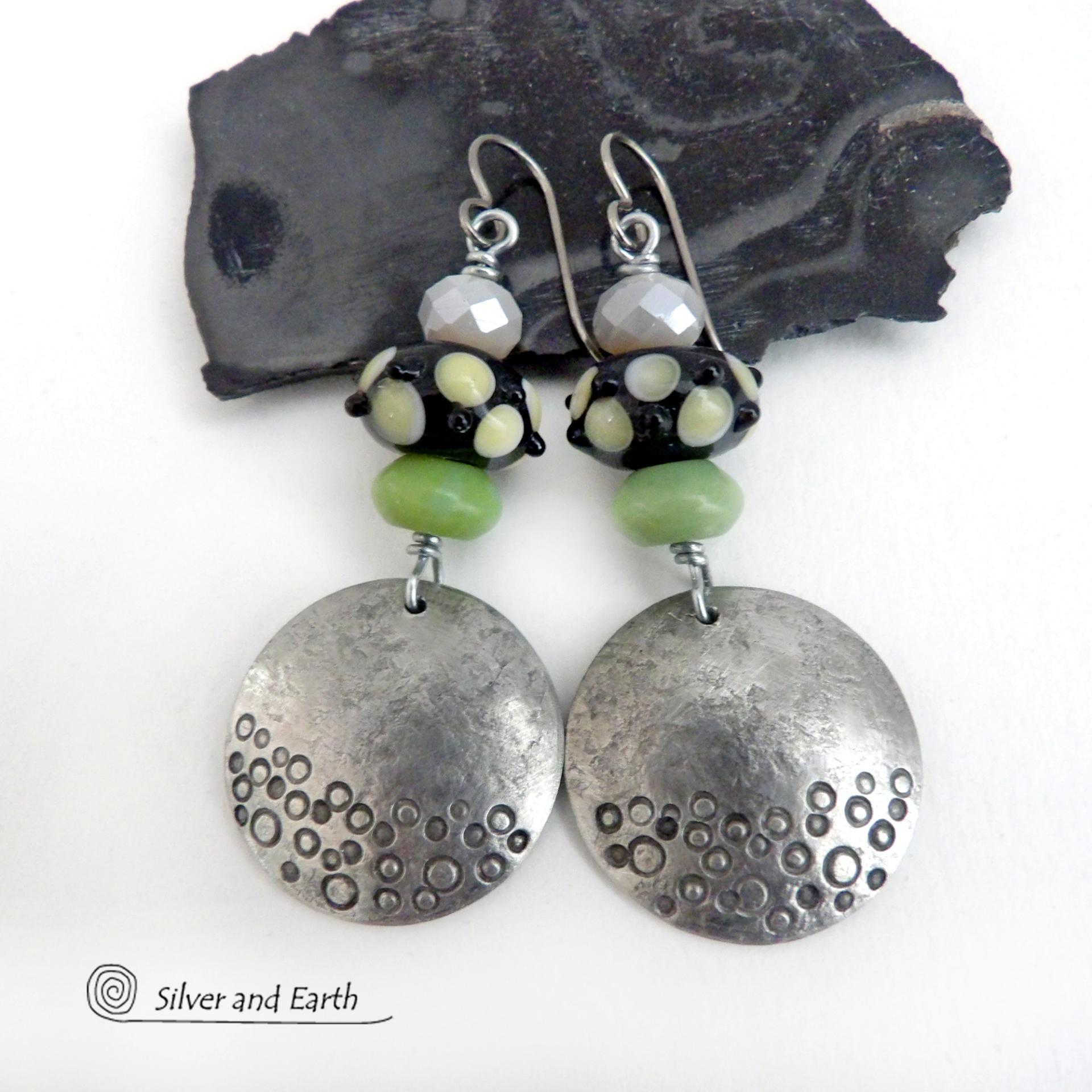 These handcrafted silver pewter earrings have a hand stamped texture and are accented with artisan handmade lampwork beads, green serpentine gemstones and off-white faceted crystal beads. The lampwork beads have a black base with off-white dots that have subtle pale yellow undertones, along with funky black bumps sticking out. Handmade in the USA by Silver and Earth. 