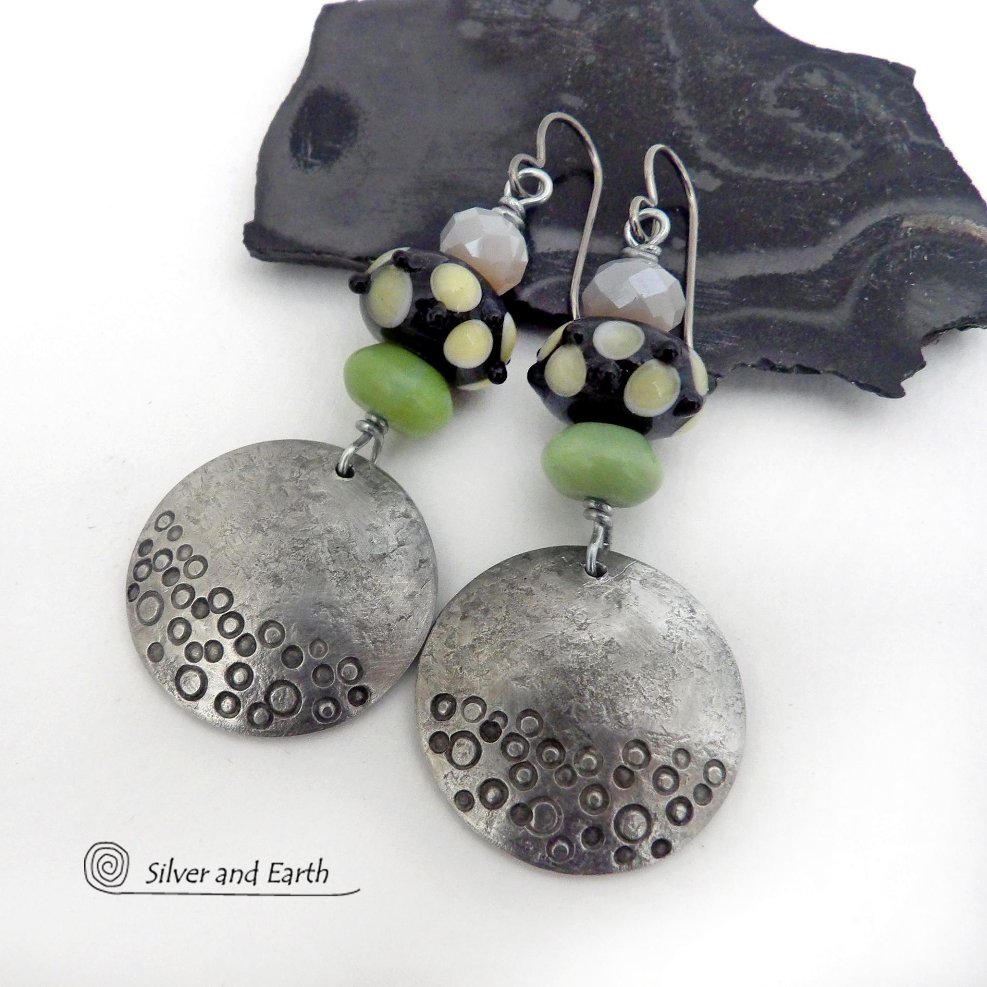 These handcrafted silver pewter earrings have a hand stamped texture and are accented with artisan handmade lampwork beads, green serpentine gemstones and off-white faceted crystal beads. The lampwork beads have a black base with off-white dots that have subtle pale yellow undertones, along with funky black bumps sticking out. Handmade in the USA by Silver and Earth. 