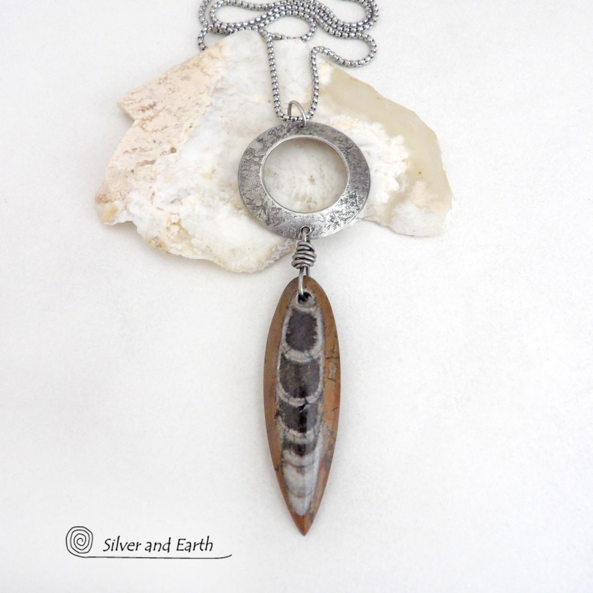 This one of a kind necklace features a natural Orthoceras fossil stone dangling from a silver pewter circle with a lightly hammered texture. This particular fossil has a brown hue - which is unusual, because these fossils are typically found in more black tones. This type of fossil is found in the Sahara Desert of Morocco where they were once ocean dwellers. Their shells were preserved in the marble in which they are found today. The design is unisex -- so it can be worn by either a man or a woman. Makes a 