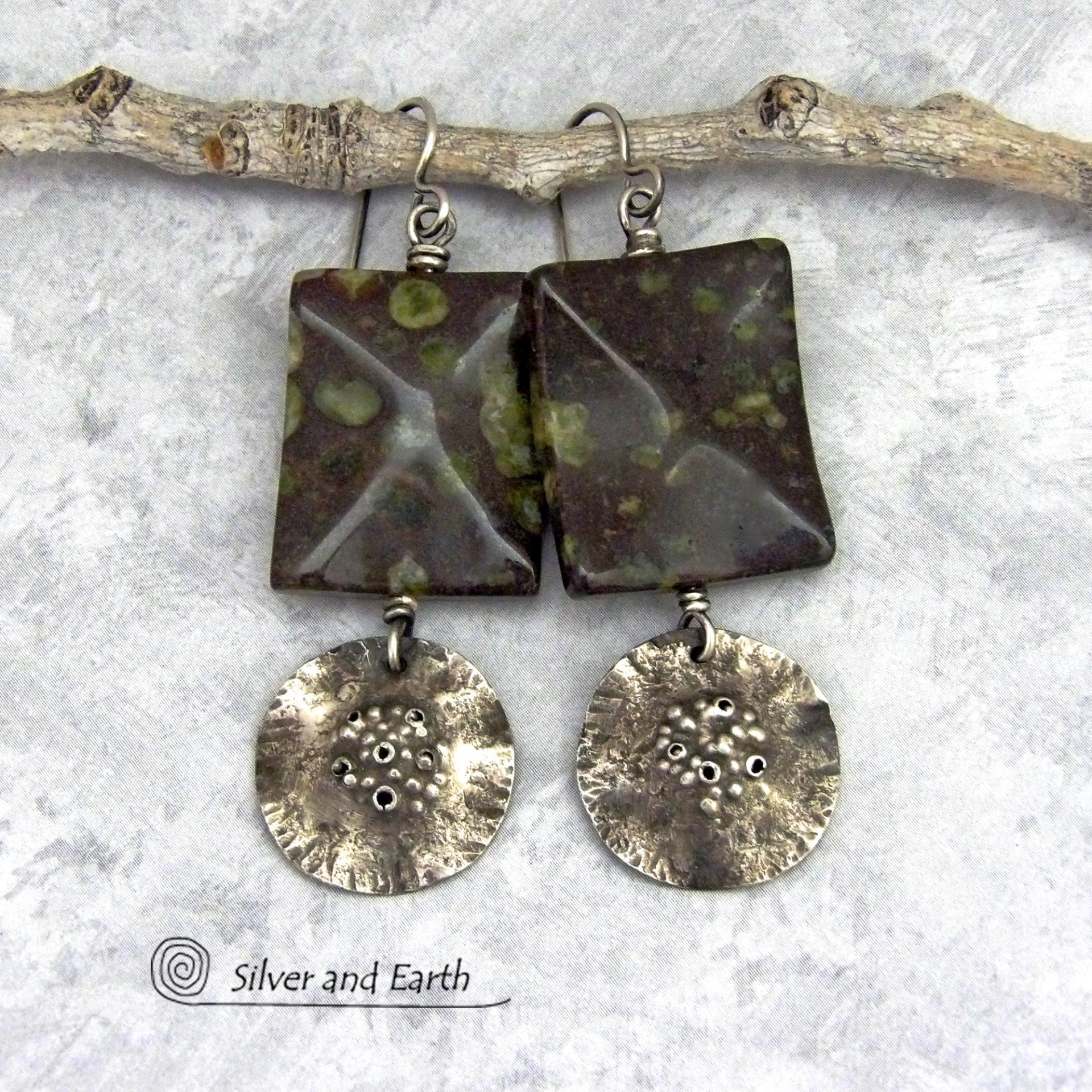 Large chunky spotted jasper stones are accented with handcrafted rustic sterling silver dangles in these one-of-a-kind earrings. The silver has an earthy hammered organic texture with a few holes for a primal effect. The stones have a dark brownish-black base with a natural matrix of green spots and a faceted finish.