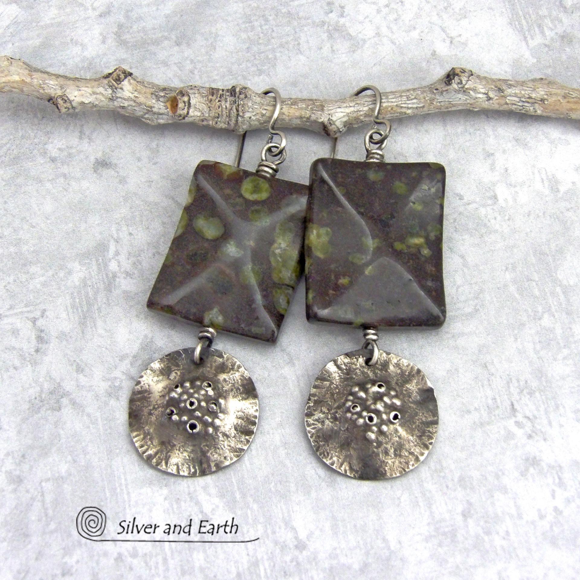 Large chunky spotted jasper stones are accented with handcrafted rustic sterling silver dangles in these one-of-a-kind earrings. The silver has an earthy hammered organic texture with a few holes for a primal effect. The stones have a dark brownish-black base with a natural matrix of green spots and a faceted finish.